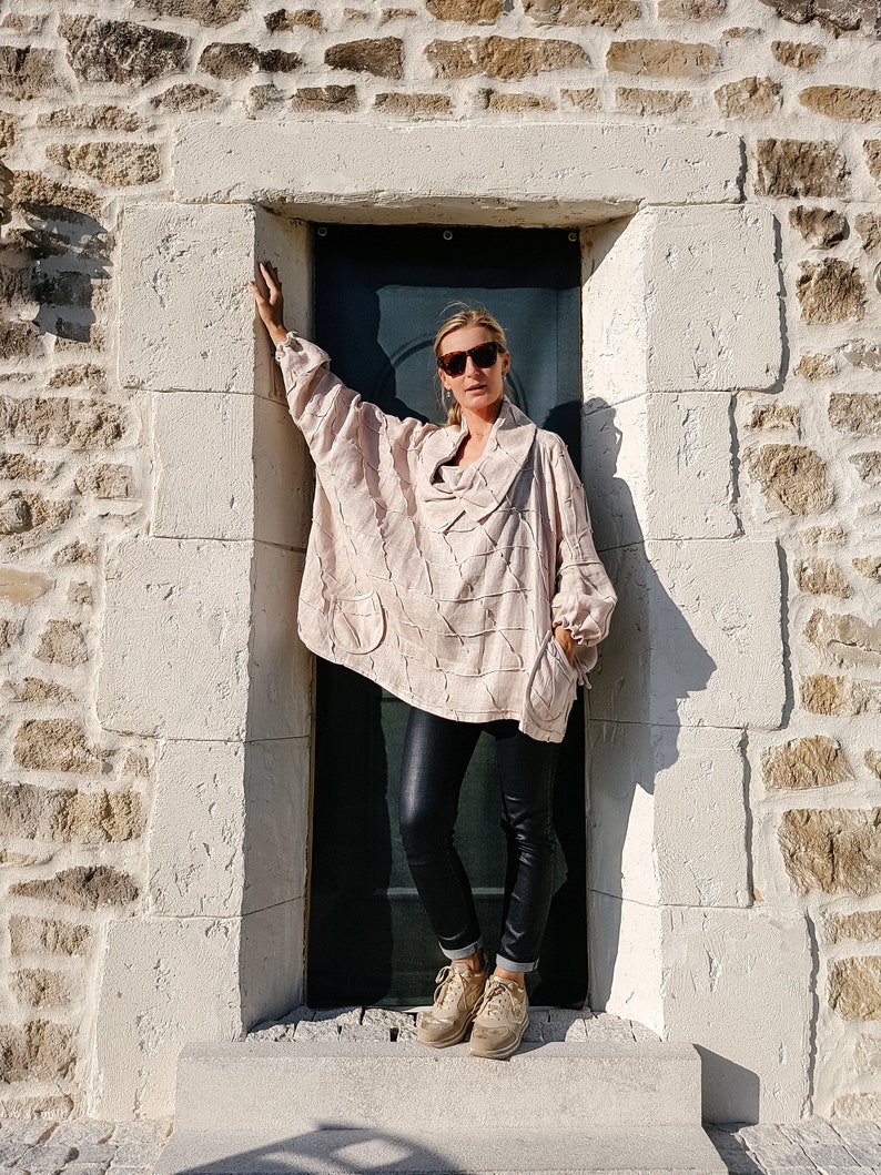 May include: A woman wearing a light pink, textured, oversized sweater with a cowl neck and black leather pants. She is standing in a doorway with a stone wall behind her. She is wearing sunglasses and brown sneakers.