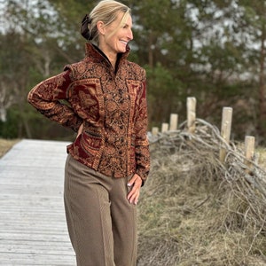 Puede incluir: Una mujer lleva una chaqueta con estampado marrón y dorado con cuello alto y un pantalón de cuadros marrones y blancos. Está de pie en una pasarela de madera con una valla en el fondo.