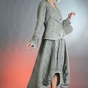 May include: A woman wearing a gray, textured, asymmetrical skirt and matching jacket. The skirt has a high waist and a flowing, pleated design. The jacket has a long, flowing sleeve and a unique, textured design. The woman is wearing red heels.