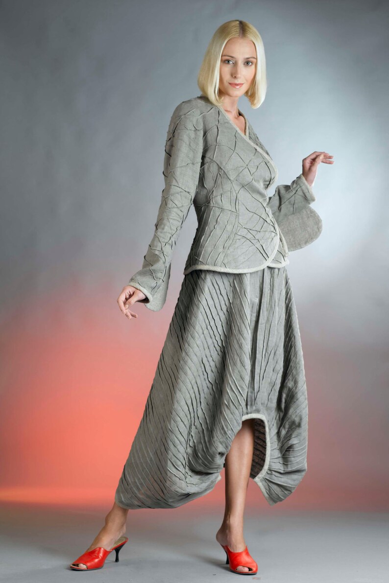 May include: A woman wearing a gray, textured, asymmetrical skirt and matching jacket. The skirt has a high waist and a flowing, pleated design. The jacket has a long, flowing sleeve and a unique, textured design. The woman is wearing red heels.