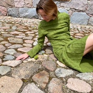 以下が含まれることがあります： 緑色のテクスチャのある長袖のドレスを着た女性が、石畳の道に座っています。
