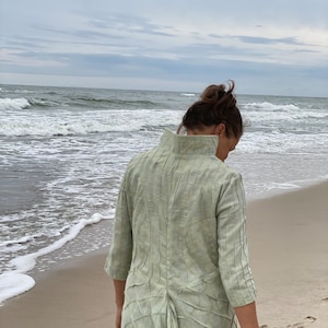 May include: A woman wearing a light green, textured, long-sleeved dress walks on a sandy beach with the ocean in the background.