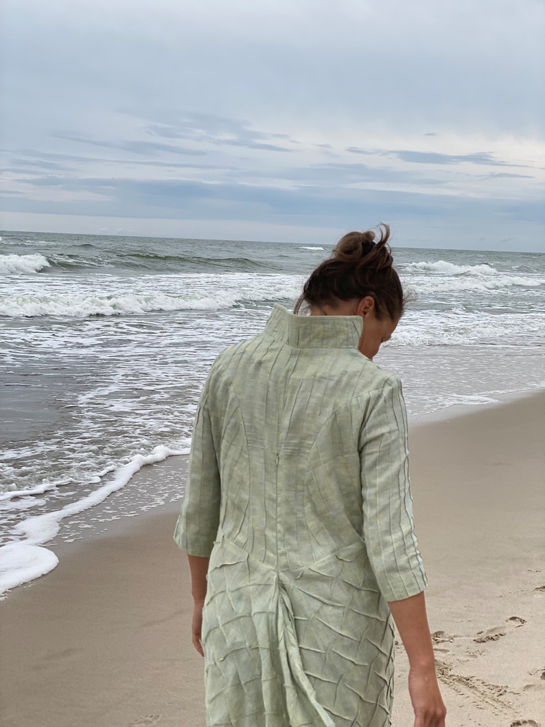 May include: A woman wearing a light green, textured, long-sleeved dress walks on a sandy beach with the ocean in the background.