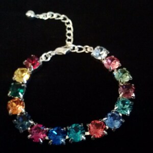 Può includere: Un braccialetto argentato con una serie di pietre preziose quadrate e colorate. Le pietre sono disposte in un motivo circolare, mostrando uno spettro di colori tra cui rosso, giallo, verde, blu e rosa. Il braccialetto ha una catena regolabile.