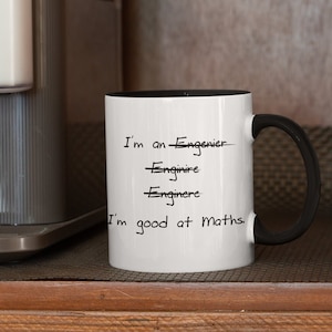 May include: White ceramic mug with a black interior and handle. The mug has the text "I'm an -Engenier, -Enginire, -Enginere, I'm good at Maths." written in black, with the incorrect spellings crossed out.