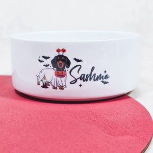 May include: White ceramic pet bowl with a Halloween-themed dachshund illustration. The dog is dressed as a ghost with a pumpkin head and bat accents. The name "Sashmio" is written in a script font.