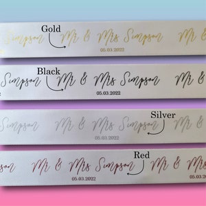 May include: White satin ribbons with gold, black, silver, and red text. The ribbons display the names "Mr & Mrs Simpson" and the date "05.03.2022" in a cursive font, ideal for wedding decorations.