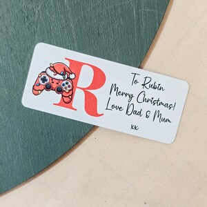 May include: A white rectangular gift tag with rounded corners. The tag features a red letter "R" and a red controller wearing a Santa hat. Text on the tag reads "To Rubin Merry Christmas! Love Dad & Mum xx".