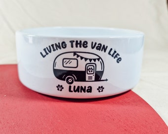 Op de afbeelding: Witte keramische voerbak voor huisdieren met een zwarte afbeelding van een camper, pootafdrukken en de naam "Luna". De tekst "Living the Van Life" staat boven de camper. De kom staat op een rood en beige oppervlak.