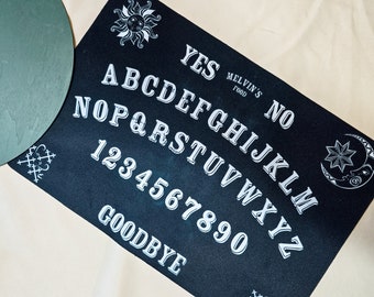 May include: A black Ouija board with white lettering and symbols. The board features the alphabet, numbers, and the words "YES", "NO", and "GOODBYE". Decorative sun and moon graphics are present. The text "MELVIN'S FOOD" is also visible.