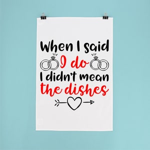 Peut inclure: Torchon blanc avec le texte "When I said I do I didn't mean the dishes" en noir et rouge. Le motif comprend deux bagues en diamant et un cœur avec une flèche.