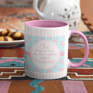 May include: White ceramic mug with a pink handle and interior. The mug features a floral and striped design with the text "Tea keeps me busy until it's acceptable to drink Prosecco."