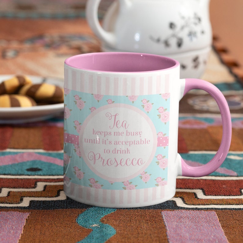 May include: White ceramic mug with a pink handle and interior. The mug features a floral and striped design with the text "Tea keeps me busy until it's acceptable to drink Prosecco."