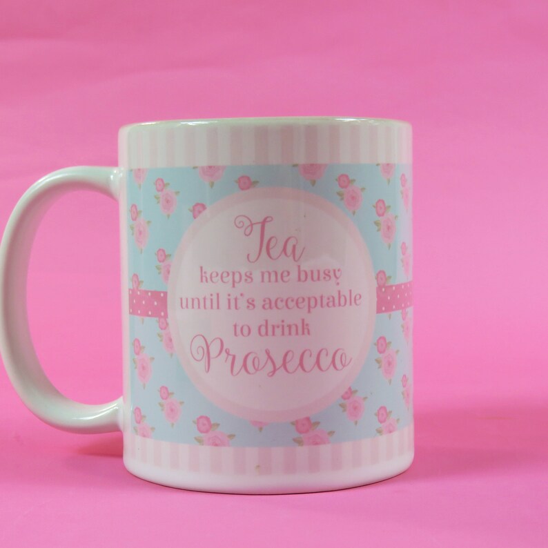 May include: White ceramic mug with a handle, featuring a light blue floral pattern and the text "Tea keeps me busy until it's acceptable to drink Prosecco" in pink script. Pink polka dot ribbon accents the design.