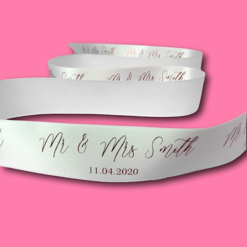 May include: White satin ribbon with the text "Mr & Mrs Smith" and the date "11.04.2020" printed in a dark color. The ribbon is curled and displayed against a pink background, suitable for weddings or special events.