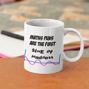 Peut inclure: Mug en céramique blanche avec l'inscription "MATHS PUNS ARE THE FIRST SINE OF Madness" en noir, avec un graphique d'onde sinusoïdale bleu et rose. La tasse a une anse incurvée et est posée sur une surface en bois.