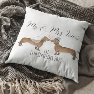 Peut inclure: Un coussin carré blanc avec un motif personnalisé. Il présente deux teckels, l'un portant un voile et l'autre un chapeau haut de forme, s'embrassant. Le texte "Mr & Mrs Jones" et "Est. 11th September 2023" sont imprimés sur le coussin.