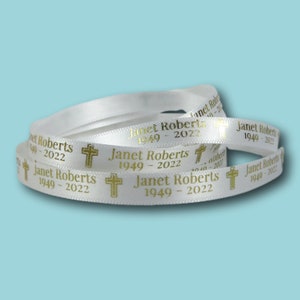 Può includere: Nastro di raso bianco con testo dorato e un motivo a croce. Il testo recita "Janet Roberts 1949-2022". Il nastro è arrotolato e si trova su uno sfondo azzurro.