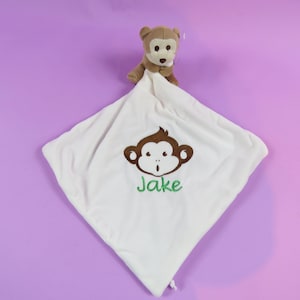 May include: A white baby comfort blanket with a brown monkey soft toy attached. The blanket has an embroidered brown monkey face and the name "Jake" in green. The background is a solid purple.