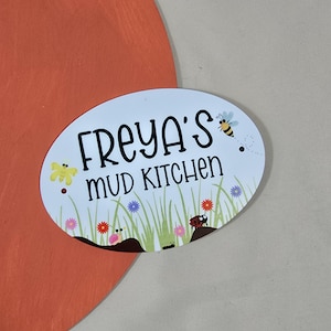 Könnte beinhalten: Ovales Schild mit dem Text "FREYA'S MUD KITCHEN" in Schwarz. Das Schild hat einen hellblauen Hintergrund mit Illustrationen einer Biene, Blumen, Gras und eines Marienkäfers. Das Schild ist vor einem orangefarbenen Hintergrund angebracht.