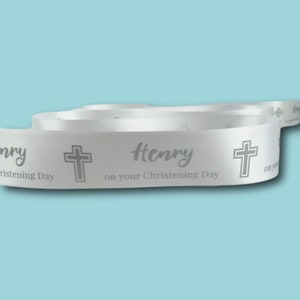 May include: White satin ribbon featuring the name "Henry", a cross, and the words "on your Christening Day" printed in grey. The ribbon is coiled against a light blue backdrop.