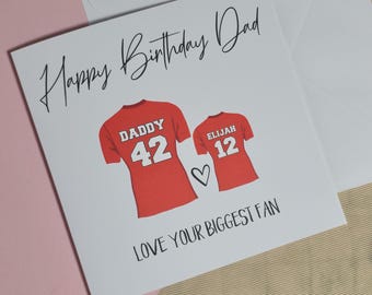 Op de afbeelding: Een witte wenskaart met de tekst "Happy Birthday Dad" in zwart schrift. De kaart toont twee rode jersey-illustraties met de namen "Daddy" en "Elijah" en de nummers "42" en "12". De tekst "LOVE YOUR BIGGEST FAN" staat onderaan.