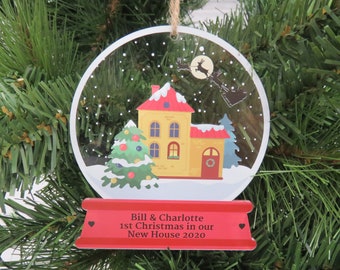 May include: A clear, round Christmas ornament with a house and Christmas tree design. The ornament has a red banner at the bottom with the text "Bill & Charlotte 1st Christmas in our New House 2020". A string is attached for hanging.