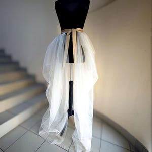 May include: A white tulle skirt with a high-low design is displayed on a black dress form. A gold-colored ribbon is tied around the waist. The skirt is long in the back and shorter in the front, creating a layered effect. The background features a staircase and a neutral-colored wall.