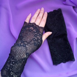 K&ouml;nnte beinhalten: Ein Paar schwarze fingerlose Handschuhe aus Spitze mit einem zarten Blumenmuster. Die Handschuhe liegen auf einem violetten Hintergrund.