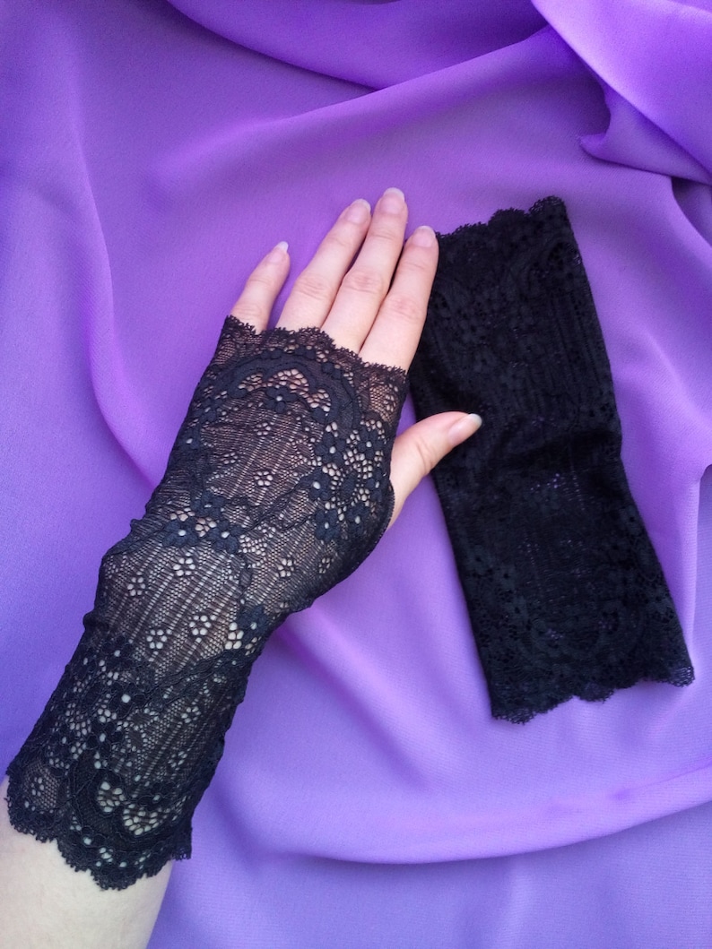 K&ouml;nnte beinhalten: Ein Paar schwarze fingerlose Handschuhe aus Spitze mit einem zarten Blumenmuster. Die Handschuhe liegen auf einem violetten Hintergrund.