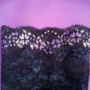May include: Close-up of black lace fabric with a scalloped edge, set against a purple background. The lace has a delicate floral pattern with hints of white and lavender. The fabric is likely used for lingerie or apparel.