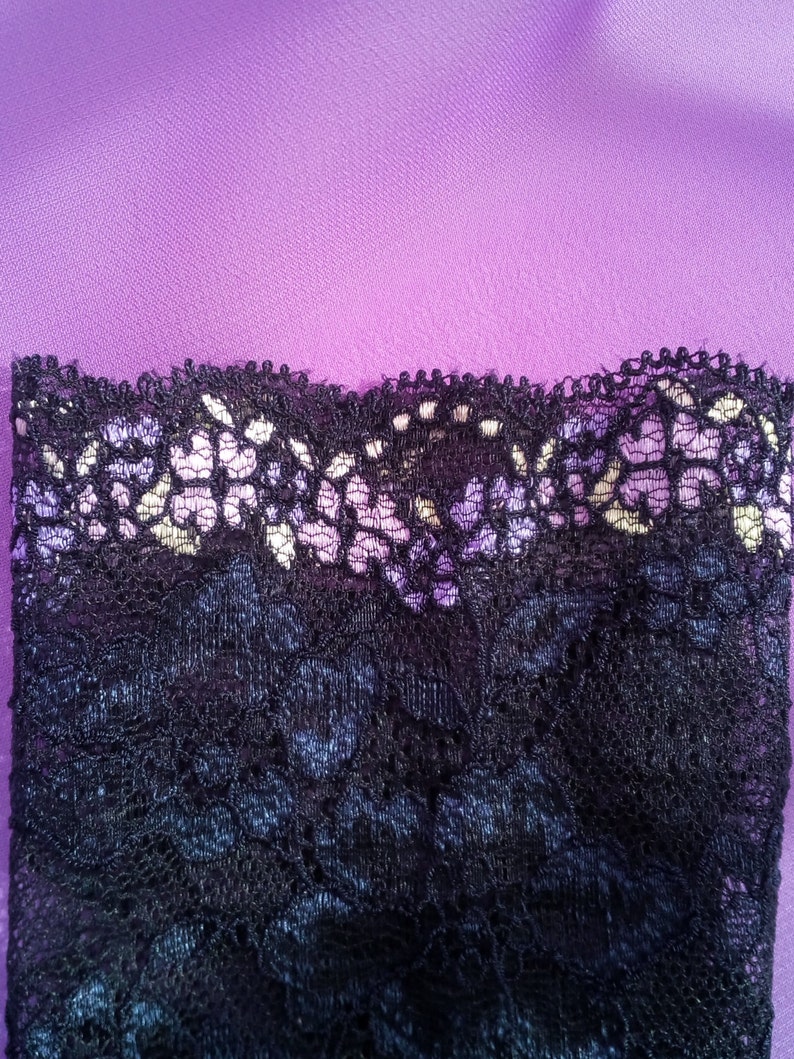 May include: Close-up of black lace fabric with a scalloped edge, set against a purple background. The lace has a delicate floral pattern with hints of white and lavender. The fabric is likely used for lingerie or apparel.