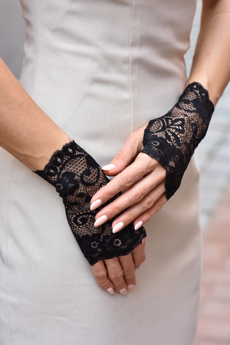Puede incluir: Un par de guantes sin dedos de encaje negro con un delicado patr&oacute;n floral. Los guantes se llevan en las manos de una persona, con las yemas de los dedos expuestas.