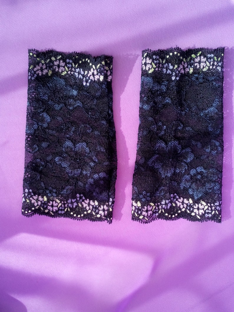 May include: Two black lace arm warmers with a floral pattern and a scalloped edge. The lace has a dark blue and purple floral design. The top edge has a white and yellow floral pattern.