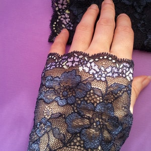 May include: Black lace fingerless gloves with a floral pattern. The gloves have a scalloped edge with a delicate floral design in shades of purple, white, and yellow. The gloves are on a purple background.