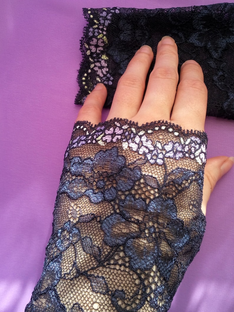 May include: Black lace fingerless gloves with a floral pattern. The gloves have a scalloped edge with a delicate floral design in shades of purple, white, and yellow. The gloves are on a purple background.