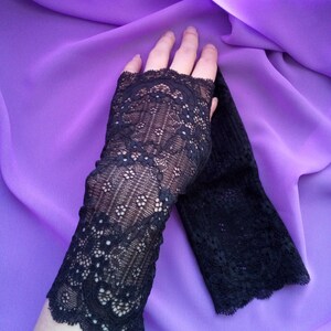 K&ouml;nnte beinhalten: Ein Paar schwarze Spitzen-Fingerhandschuhe mit einem zarten Blumenmuster. Die Handschuhe liegen auf einem violetten Stoffhintergrund.