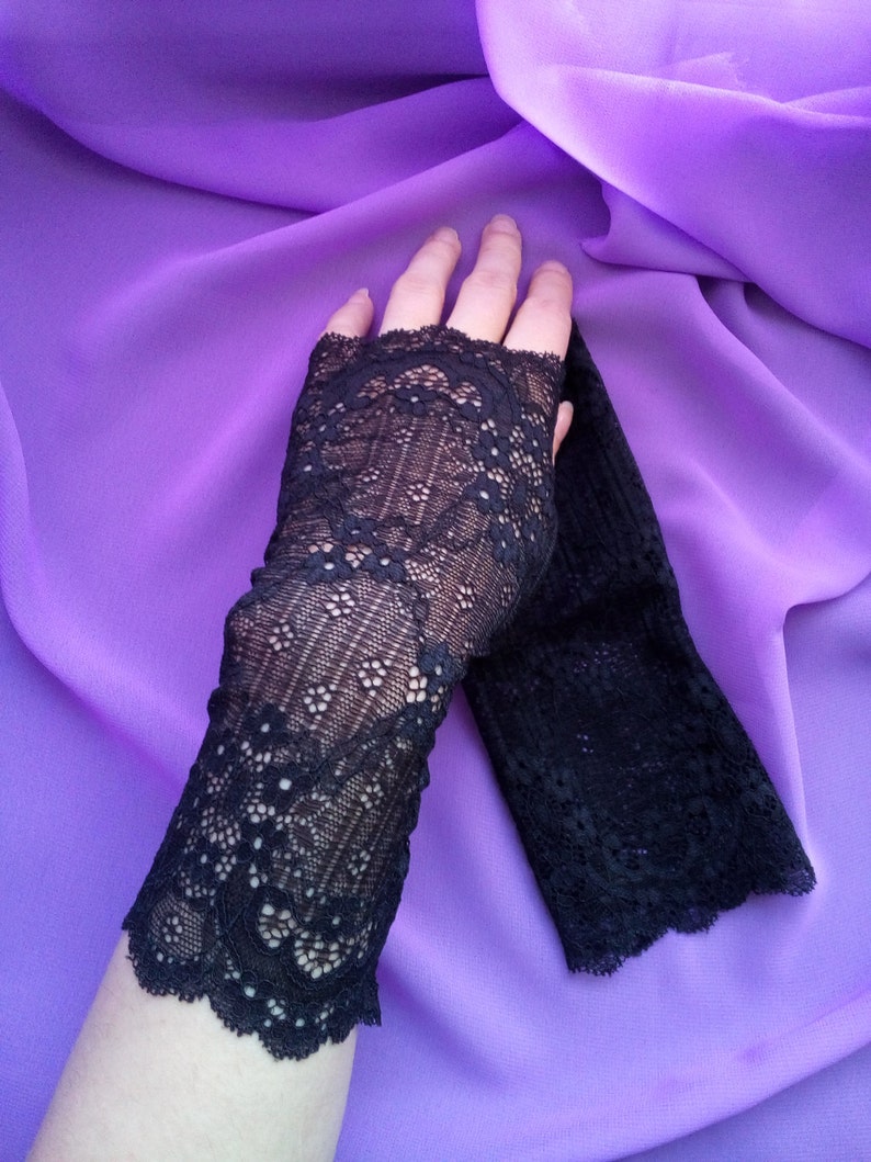 K&ouml;nnte beinhalten: Ein Paar schwarze Spitzen-Fingerhandschuhe mit einem zarten Blumenmuster. Die Handschuhe liegen auf einem violetten Stoffhintergrund.