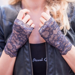May include: Dark navy blue lace fingerless gloves with a scalloped edge. The gloves are worn over a black top and a black leather jacket. The gloves have a floral pattern and a delicate, feminine design.