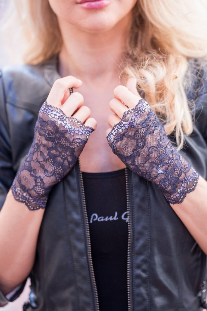 May include: Dark navy blue lace fingerless gloves with a scalloped edge. The gloves are worn over a black top and a black leather jacket. The gloves have a floral pattern and a delicate, feminine design.