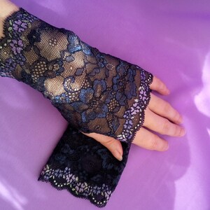 May include: Black lace fingerless gloves with a floral design and scalloped edges. The gloves feature blue and purple accents and are displayed on a purple background. The delicate lace adds a touch of elegance.