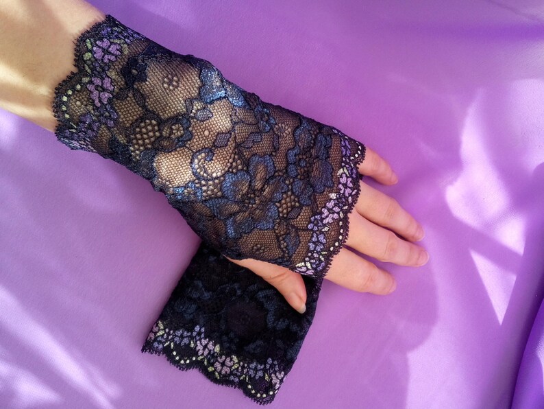 May include: Black lace fingerless gloves with a floral design and scalloped edges. The gloves feature blue and purple accents and are displayed on a purple background. The delicate lace adds a touch of elegance.