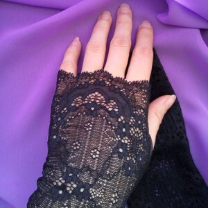 K&ouml;nnte beinhalten: Ein Paar schwarze fingerlose Handschuhe aus Spitze mit einem zarten Blumenmuster. Die Handschuhe werden auf einem violetten Hintergrund gezeigt.