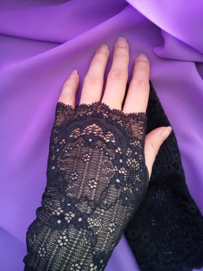 K&ouml;nnte beinhalten: Ein Paar schwarze fingerlose Handschuhe aus Spitze mit einem zarten Blumenmuster. Die Handschuhe werden auf einem violetten Hintergrund gezeigt.