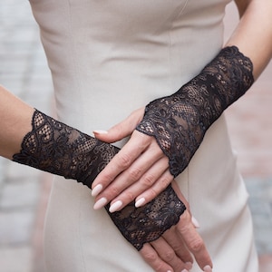 May include: A pair of black lace fingerless gloves with a delicate floral pattern. The gloves are worn on a person's hands, with the fingertips exposed.