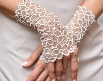 May include: Elegant white lace fingerless gloves with a floral pattern. The gloves are worn over a neutral-colored dress. The intricate design and delicate fabric create a sophisticated look, perfect for special occasions.