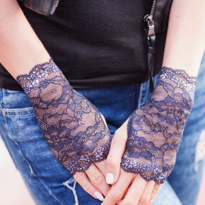 May include: Close-up of a pair of dark blue lace fingerless gloves. The gloves have a delicate floral pattern and a scalloped edge. The person is wearing a black top and blue jeans.