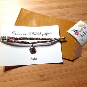 Op de afbeelding: Een armband met bloemenpatroon en een lieveheersbeestjesbedel, gepresenteerd op een witte kaart. De kaart heeft Franse tekst en de naam "Jules" onder de armband. Er is ook een klein cadeaulabel met bloemmotieven.