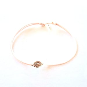 May include: A light pink cord bracelet featuring a small, oval-shaped gold-toned metal charm. The charm displays a detailed embossed figure on its surface. The bracelet is secured with adjustable sliding knots, allowing for a flexible fit on the wrist.
