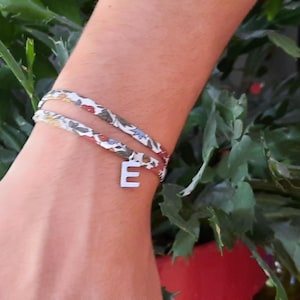 Peut inclure: Un bracelet en tissu à double tour orné d'un motif floral dans des tons de rouge, jaune et gris. Le bracelet est agrémenté d'une breloque en métal argenté en forme de lettre majuscule E, suspendue directement au ruban de tissu.